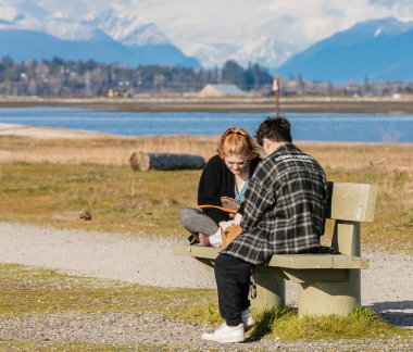 Güneşli bir sonbahar gününde parkta bankta oturan genç bir çift. Gençler, Kanada 'da 8 Nisan 2021 tarihli White Rock BC parkında piknik yapıyorlar. Sokak görünümü, seçici odak, metin için telif alanı