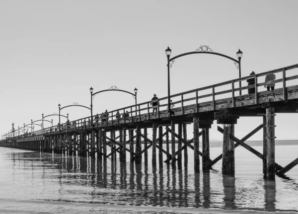 Beyaz Kaya 'daki ahşap iskele, BC çapraz olarak görüntüye yayılıyor. Parıldayan ışıklar denize yansıyor. Siyah ve beyaz görüntü: Gün batımında sahil ve iskele, Surrey, BC, Kanada-22 Şubat 2022