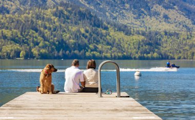 Köpekli bir çift gölde rıhtımda oturuyor. Kanada 'daki Cultus Gölü BC' de köpeklerle iskelede oturan romantik bir çiftin arka görüntüsü. Seyahat fotoğrafı, seçici odak