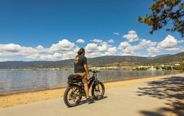 Bisikletli bir adam yaz tatili için sahilde dinleniyor. Yaz Konseptinde spor etkinliği ve eğlence. Bisikletçi, plajda duruyor. Okanagan Gölü, BC Kanada-6 Haziran 2022.