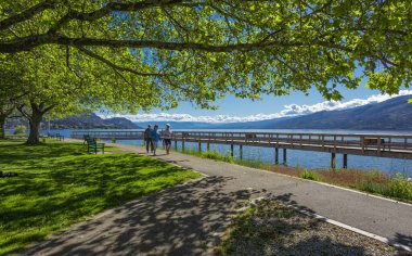 İnsanlar yaz parkında yürüyor. Güneşli bir sabahta parkta güzel bir ara sokak. Yeşil bir parkta yürüyen yaşlı insanlar. Peachland BC Kanada-Haziran 7. 2022-Seyahat fotoğrafı, aktif yaşlanma, yaz tatili konsepti