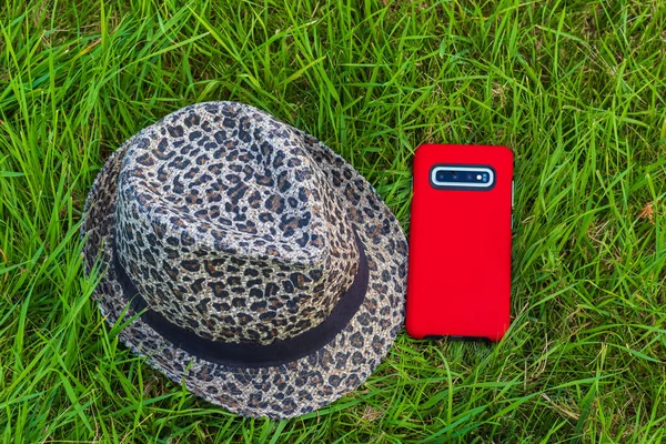 Mobile smart phone in red cover and hat on green grass. Smartphone in red case on green grass. Top view, nobody, natural background