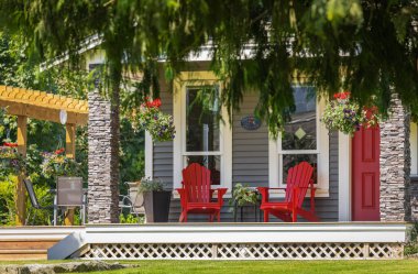 View of a large front porch with furniture and potted plants. Rustic, country style front porch seating with red Adirondack chairs and fresh flowers. Nobody,