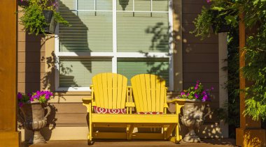 View of a large front porch with furniture and potted plants. Rustic, country style front porch seating with yellow Adirondack chairs and fresh flowers. Nobody,