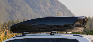 Car with the roof rack with cargo box. Black plastic car rooftop cargo box or roof carrier for traveling. Removable storage container mounted on car roof rack. Beautiful nature background