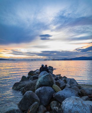 Sunset scenic over the beach. Young couple sitting on the rocks enjoying the beautiful sunset at the Pacific ocean Canada. Travel photo, selective focus, blurred, copy space for text