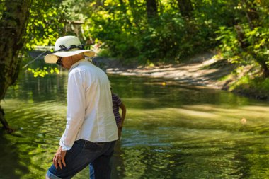 Yaşlı bir kadın yaz şapkası takıyor ve parkta su kenarında dinleniyor. Aktif yaşlanma, emeklilik sonrası yaşam, bulanık geçmiş, seyahat fotoğrafı, seçici odak - 24 Ağustos 2022-Surrey BC Kanada