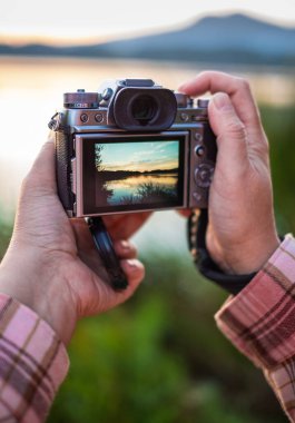 Aynasız bir dijital fotoğraf makinesi tutarak manzara fotoğrafı çekmeye hazırlanın. Kamerasız günbatımının fotoğrafını çeken eller. El kameraları güneş ışığıyla çekim yapıyor. Bulanık,