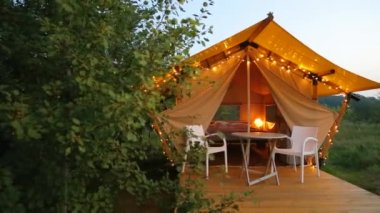 Glamping terrace in the evening forest. Cute fabric tents with evening illumination in a forest environment in an eco camp. Budget glamping for family outdoor recreation.