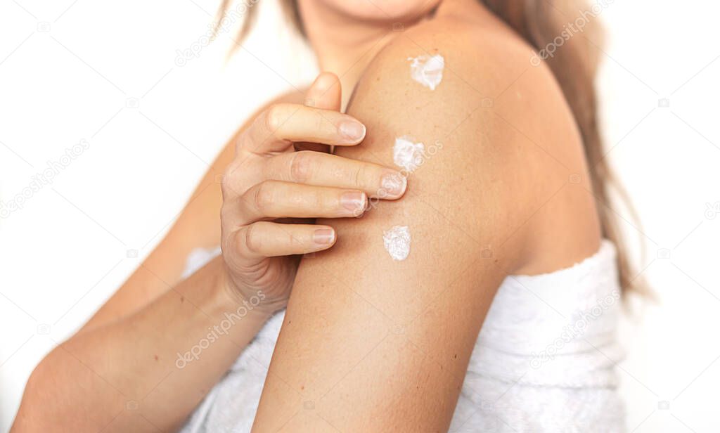 Closeup of young woman wrapped in towel applying body cream or lotion on shoulder after shower