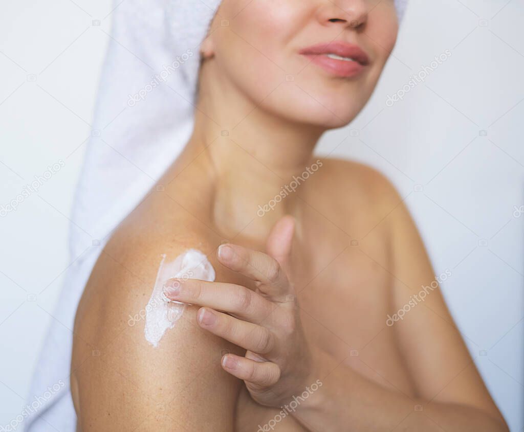 Closeup of young woman wrapped in towel applying body cream or lotion on shoulder after shower.