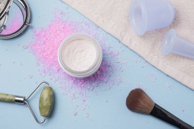 Kremalı düz yatak, banyo gülü tuzu, masaj tuzu, blu arka planda süpürge kavanozları. Spa ve doğal cilt bakımı kavramı.
