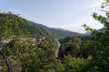 Mavi suyu olan bir nehir manzarası, solda kırmızı çatılar ve ön planda yeşil ağaçlar ve çalılar, uzaklarda mavi gökyüzü olan güzel dağ manzarası