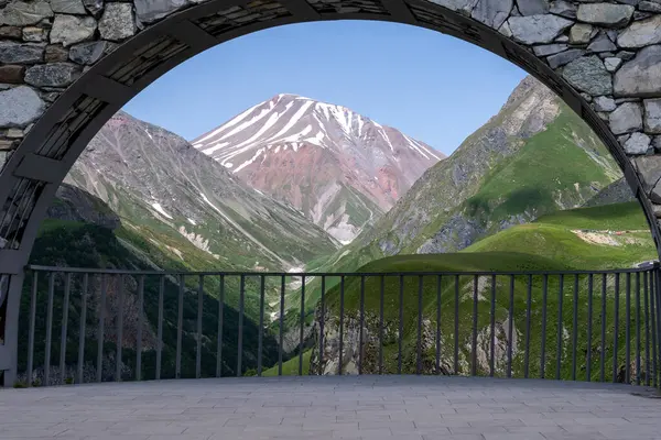 Arkasında mavi gökyüzü olan güzel bir Gürcistan dağını görebileceğiniz metal çitli büyük taş kemer.