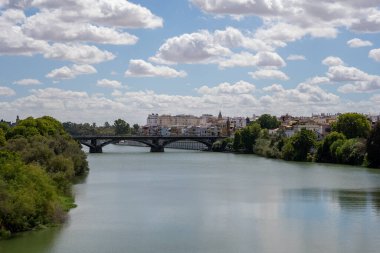 İspanya 'nın Seville şehrinde akan büyük, geniş, yeşil bir nehir. Her iki tarafında yeşil ağaçlar ve beyaz evler olan ve uzaktaki eski bir taş köprü.