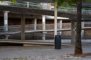 Kenarlarında metal korkuluklar olan zigzag çizgileri oluşturan bir rampanın olduğu park ve ön planda atık kutusu olan ahşap bir sandık var.
