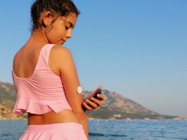Girl checks glucose level  with CGM device before enters the sea. On the right arm is placed white sensor witch send information to the CGM device. Swimming with this type of sensor is possible. Copy space for your text on the right side. clipart