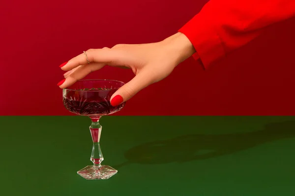 Vánoční novoroční kreativní kompozice s dámskou rukou a sklenkou červeného šampaňského na červeném a zeleném pozadí — Stock fotografie