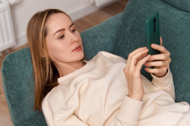 Genç yetişkin bir kadın, akıllı telefonuyla kanepede çalışıyor. Ev kavramından çalış