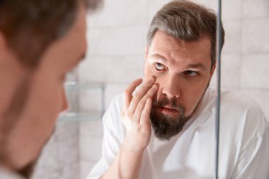 Portrait of man in white tshirt looking at the eye in the mirror. Healthcare concept.