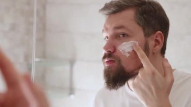 Beyaz tişörtlü bir adamın portresi banyoda yüzüne krem sürüyordu. Güzellik tedavi konsepti