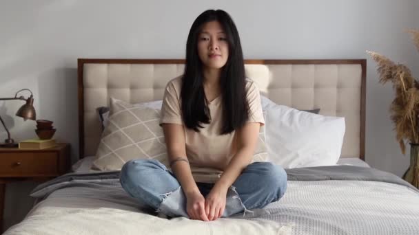 Vonzó mosolygós ázsiai nő alkalmi ruhák ül az ágyon otthon — Stock videók