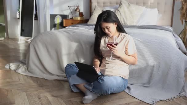 Счастливая красивая женщина в повседневной одежде держа бокал вина работает на цифровой планшет сидя возле кровати — стоковое видео
