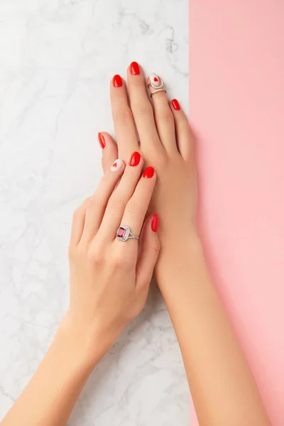 Manicured womans hands with rings on pink and marble background. Fashionable red nail design with heart — Foto Stock