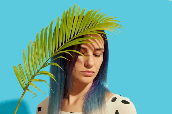 Young beautiful woman with blue hair holding palm leaf on bright background — стоковое фото