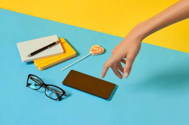 Akıllı telefon ve kırtasiyeli manikürlü kadın eli mavi masanın üzerinde. Okul işleri konseptine geri dönelim..