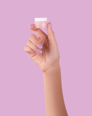 Womans hand holding jar with cream on pastel lavender background. Beauty wellness skincare concept.