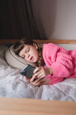 Sevimli küçük bebek pembe hırkalı ve uzun saçlı beş yaşında kız sıkılıyor ve akıllı telefondan mesaj atıyor ve yastığa uzanıyor.