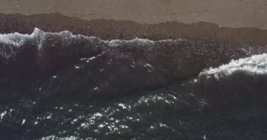 4K yukarıdan aşağıya güzel bir plaj anteni- yukarıdan siyah kumlarla dolu boş bir volkanik plaja çarpan okyanus dalgalarının kuş bakışı görüntüsü.