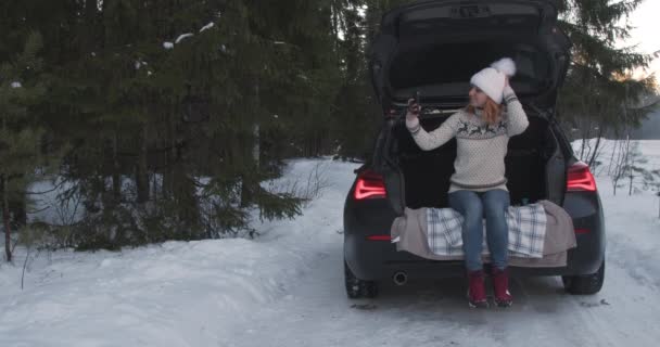 電話で自撮りし 冬の雪上車のトランクの中で美しい冬の天気を楽しむ若い女性 — ストック動画