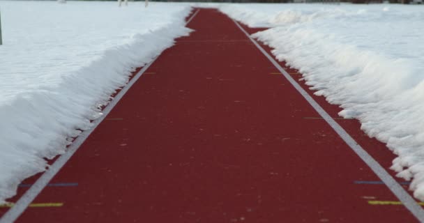 屋外スポーツスタジアムでのランニングトラックのクローズアップ ローアングルショット 雪に囲まれた白い線のマーカーを持つ赤いレーストラック — ストック動画