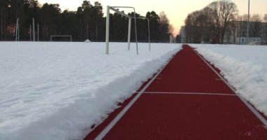 Açık hava spor stadyumundaki bir koşu pistinin alçak açılı çekimi. Beyaz çizgili ve karla çevrili kırmızı bir yarış pisti 