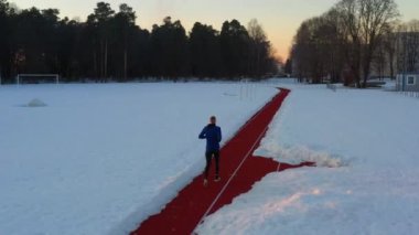 Beyaz tenli genç bir erkek atlet, gün batımında karla çevrili atletizm stadyumunda yarış pistinde koşuyor.
