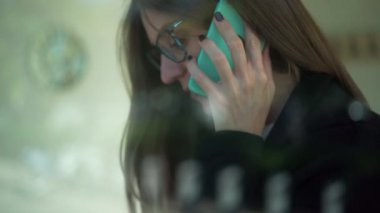 Camdan, gözlüklü, uzun düz saçlı ve resmi giyinmiş bir kadın kahve dükkanında otururken akıllı telefondan konuşuyor.