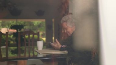 Sıcak giysiler içinde gülümseyen yaşlı bir kadın tablet ve çay bardağıyla masaya oturmuş ekrana bakıyor.