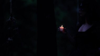 Yanan mumla esrarengiz cadı gecenin karanlığında karanlık ormanda yürüyor ve ayinler için ahşap sihirli aksesuarlara dokunuyor.