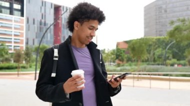 Cheerful young black male millennial with Afro hair in trendy outfit standing on city street with takeaway coffee and reading message on smartphone