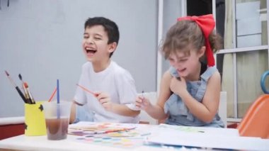Masada renkli suluboya boyalı boya fırçaları ve beyaz tahtalı malzemelerle dolu kağıtlar taşıyan pozitif çocuklar.