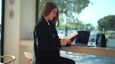 Gözlüklü kadın girişimcinin kafede dizüstü bilgisayarla çalışırken planlamacıdan not alışının yan görüntüsü