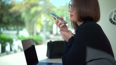 Uzun düz saçlı, gözlüklü, resmi bir kadın kahve dükkanında otururken sesli mesaj kaydediyor ve ajandasına notlar yazıyor.