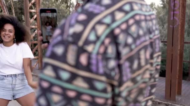 ゆりかごアフリカ系アメリカ人女性とともにスマートフォン撮影ビデオの遊び心のあるガールフレンドダンスオンブリッジ — ストック動画
