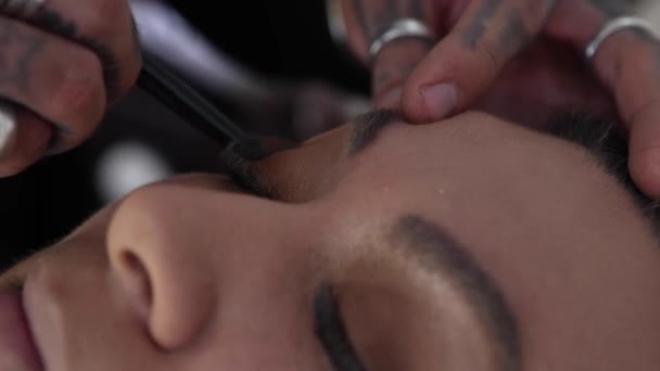 在专业化妆品工作室工作期间 戴面具的匿名男子在金发女人脸上抹黑的基础 — 图库视频影像