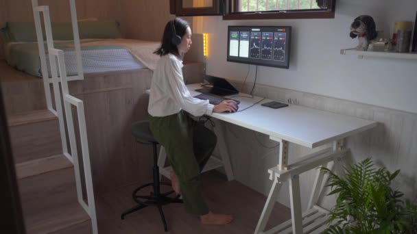便利な職場での暗号通貨の価値の変化の動的を示すチャートでコンピュータ上で働くヘッドフォンで集中アジアの女性 — ストック動画