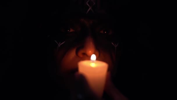 カメラを見て 暗闇の中で炎を吹いている絵の顔の近くに燃えるろうそくと魔女 — ストック動画