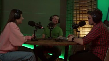 Modern stüdyoda podcast yaparken mikrofonlu masada oturan ve iletişim kuran gündelik kıyafet ve kulaklıklar takan konsantre genç erkek ve bayan meslektaşlar.
