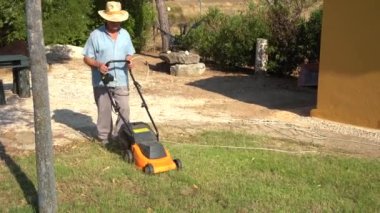 Full body of male gardener in hat mowing grassy lawn near bushes and trees in summer